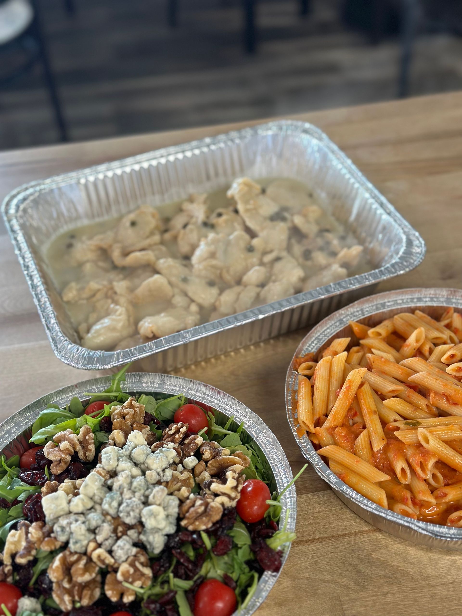 Three foil containers of food on a table: chicken in sauce, pasta in red sauce, and salad with walnuts and cheese.