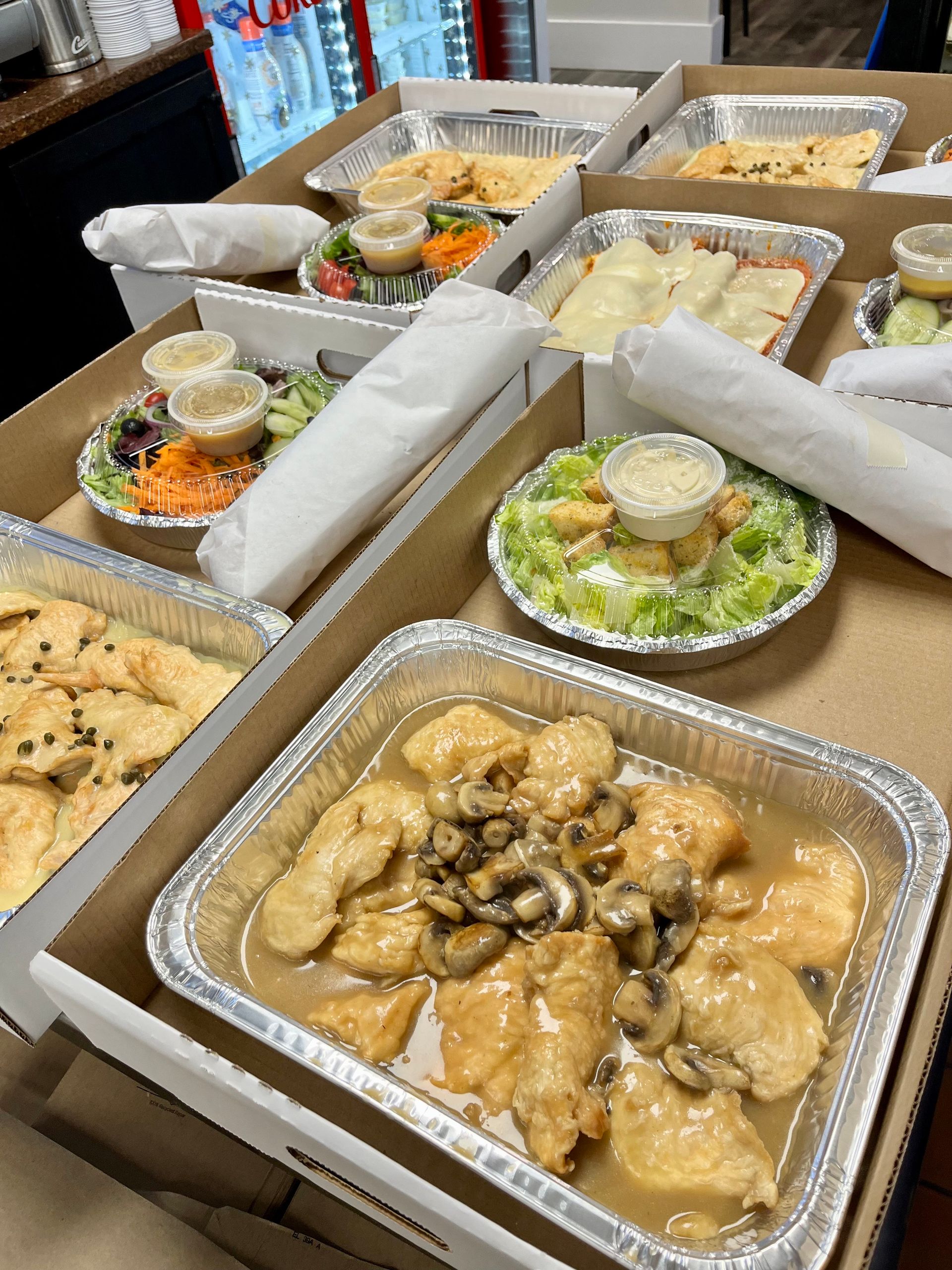 Several takeout meals in aluminum containers, including salads, pasta, and chicken with sauce.