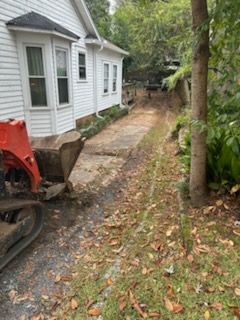 Excavator next to a white house, working on a narrow driveway lined with trees and foliage.