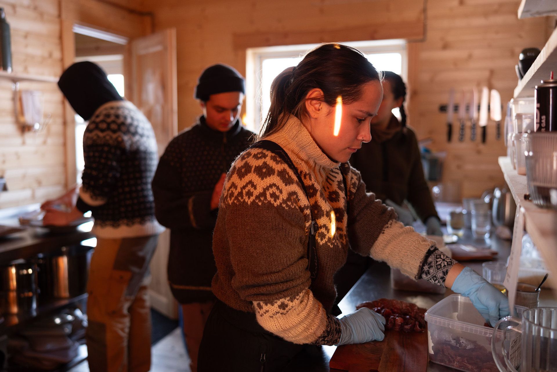 Mennesker i et trærum tilbereder mad. Kvinde i sweater og handsker er fokuseret på opgaven.