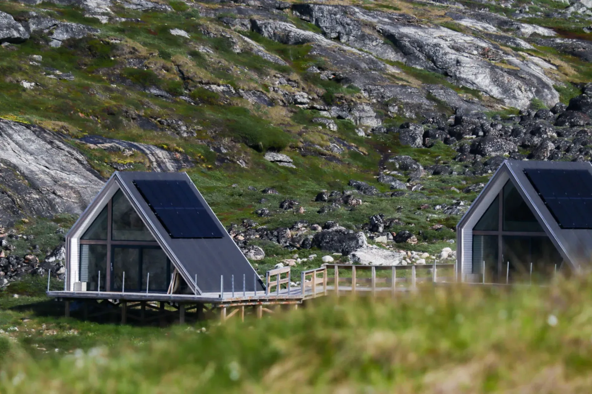 To moderne hytter med solpaneler på skrånende tage, beliggende op ad en klippefyldt bjergskråning og grøn vegetation.