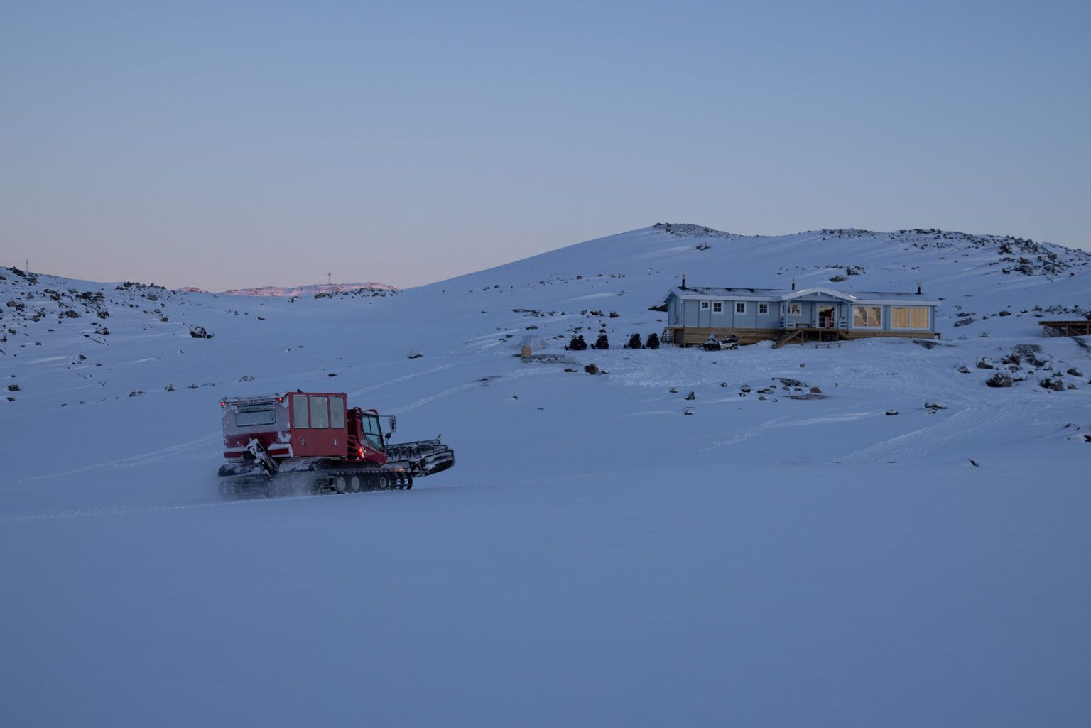 Snowcat-køretøjet kører hen over et snedækket landskab mod en bygning på en bakke under en tusmørkehimmel.