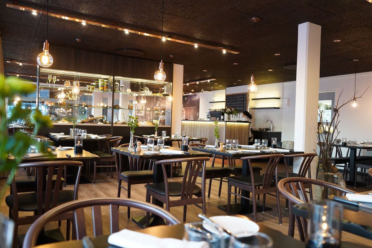 Restaurantinteriør: borde dækket med glas, mørke træstole, lyst trægulv, sort loft.