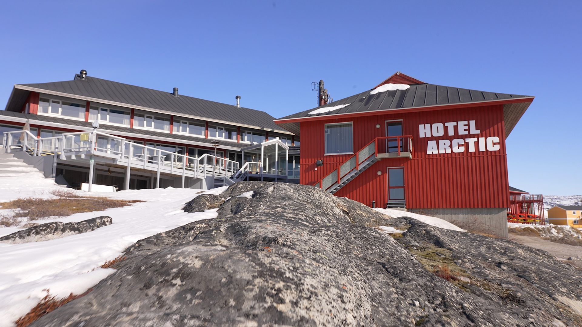 Hotel Arctic i røde og grå bygninger, delvist på snedækkede klipper, mod en blå himmel.