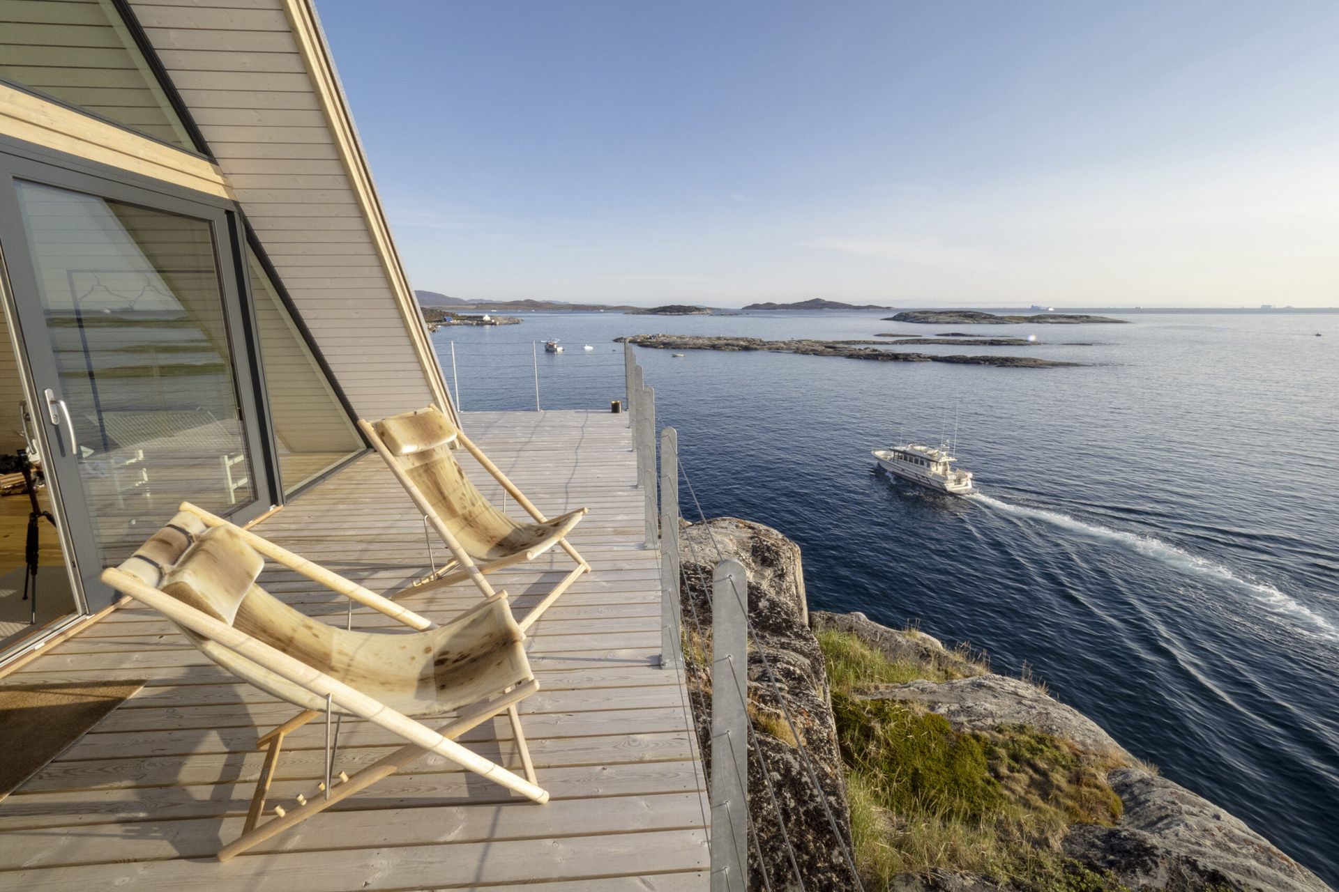 To strandstole på en terrasse med udsigt over vandet, med en båd der passerer og en moderne kahyt.