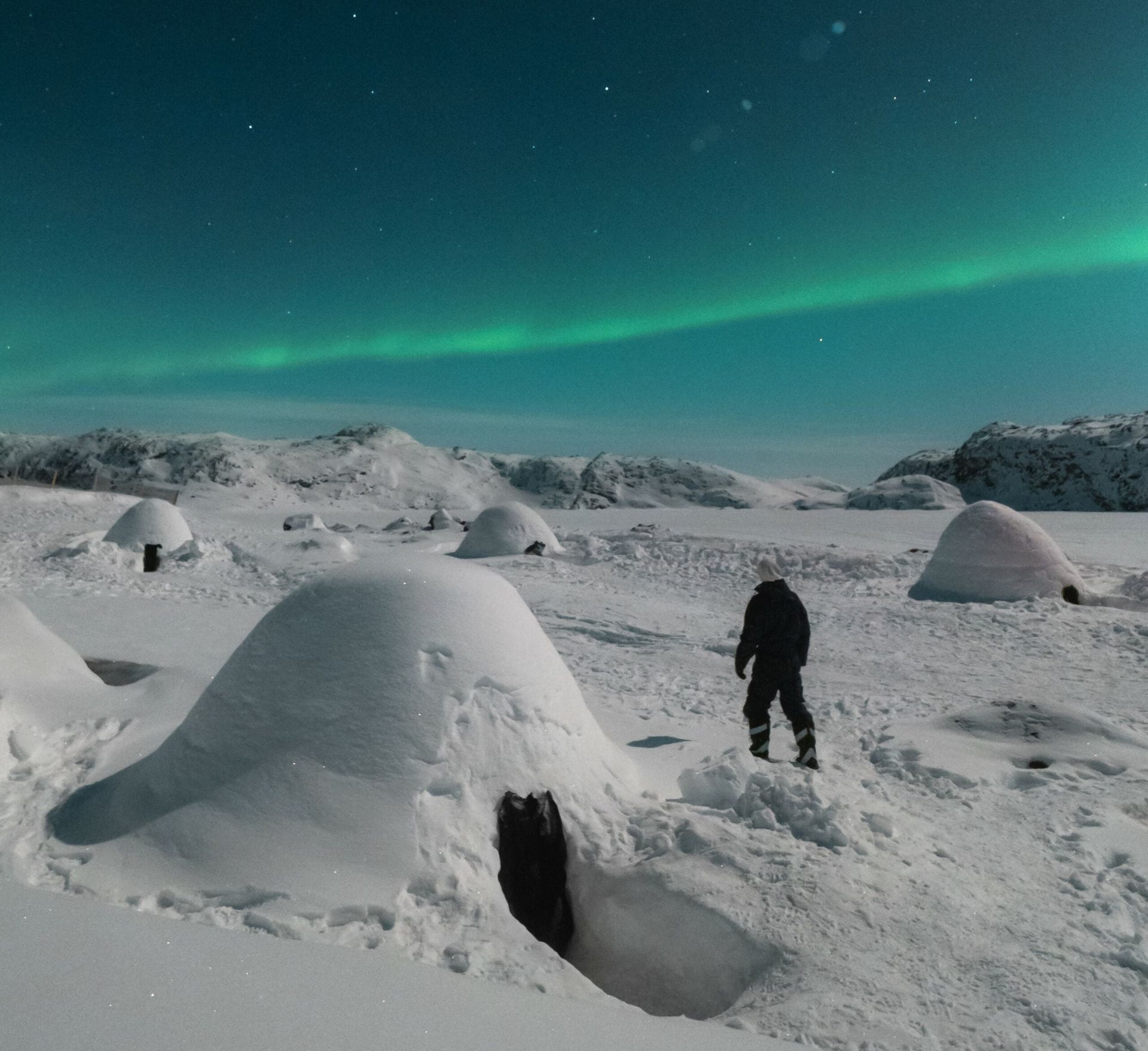 En person står i nærheden af ​​igloer under et grønt nordlys. Sne dækker jorden og de omkringliggende bakker.