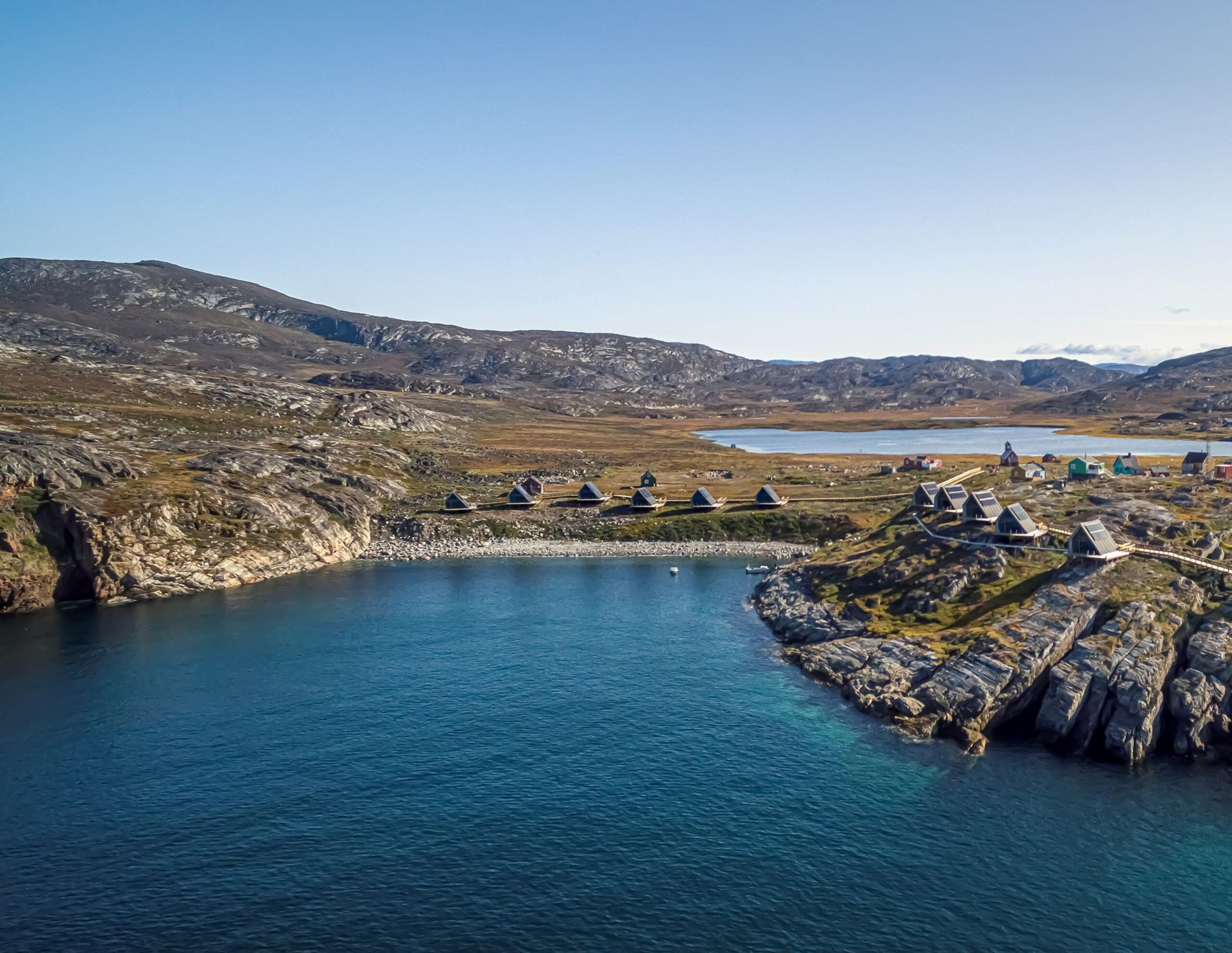 Kystlandsby i Grønland med turkisblåt vand, brunt klippefyldt terræn og blå himmel.