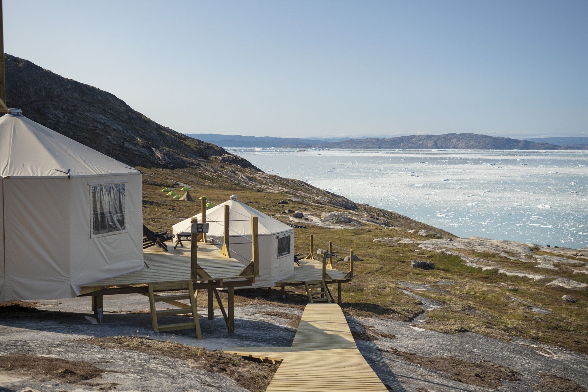 Mand trækker en slæde hen over et snedækket landskab, en gul bygning i baggrunden. Klar blå himmel.