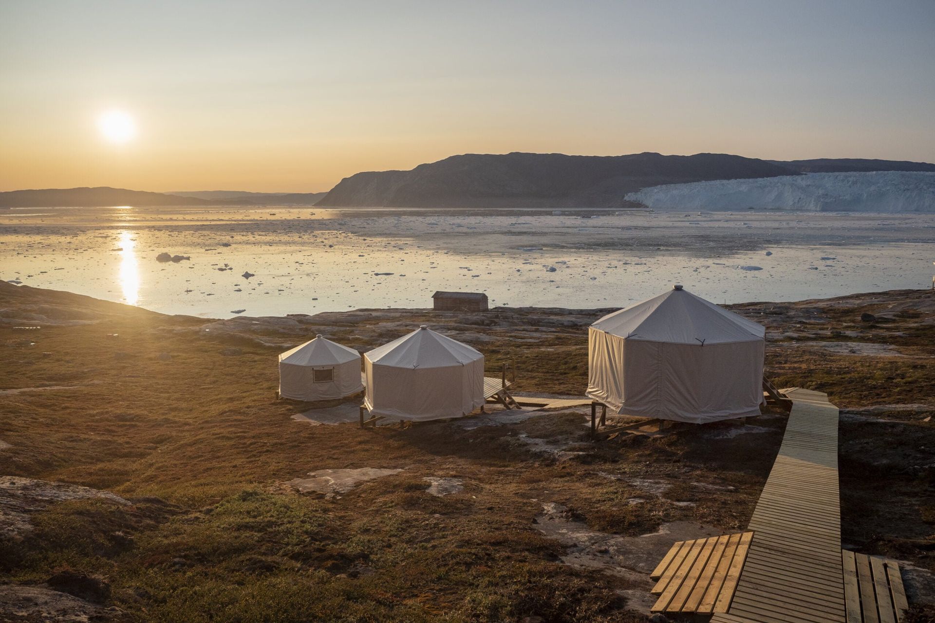 Tre hvide jurter på en græsklædt strand med et solbeskinnet hav og en gletsjer i baggrunden.