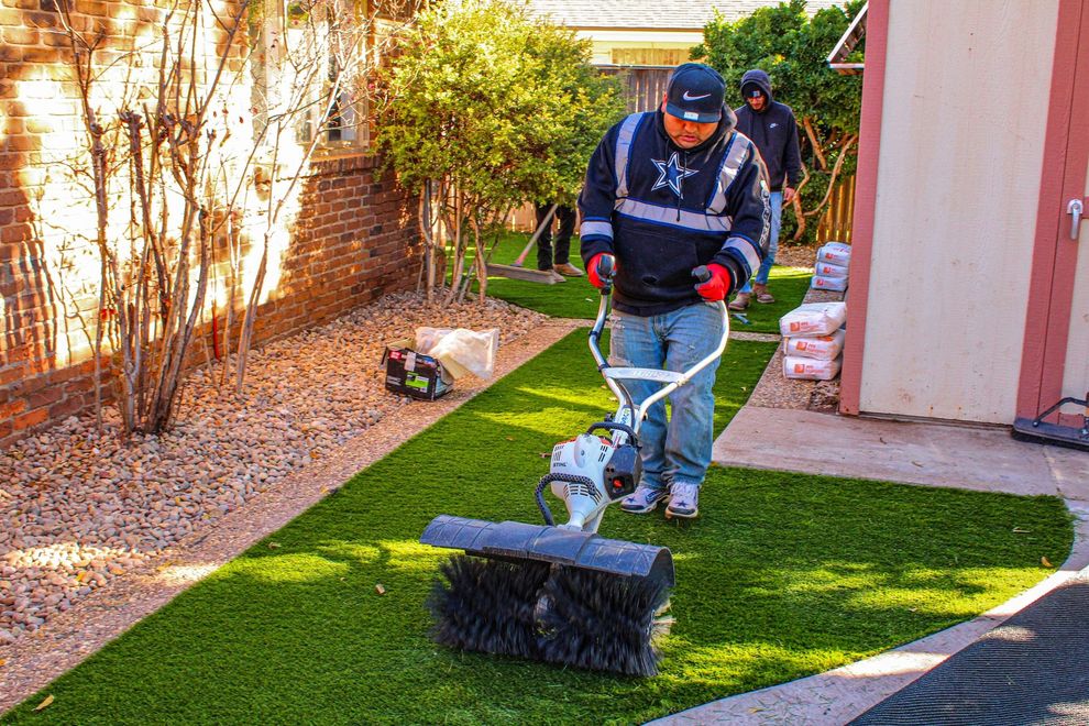 Two people in dark jackets walking a small dog on a leash across a sunny artificial grass yard.