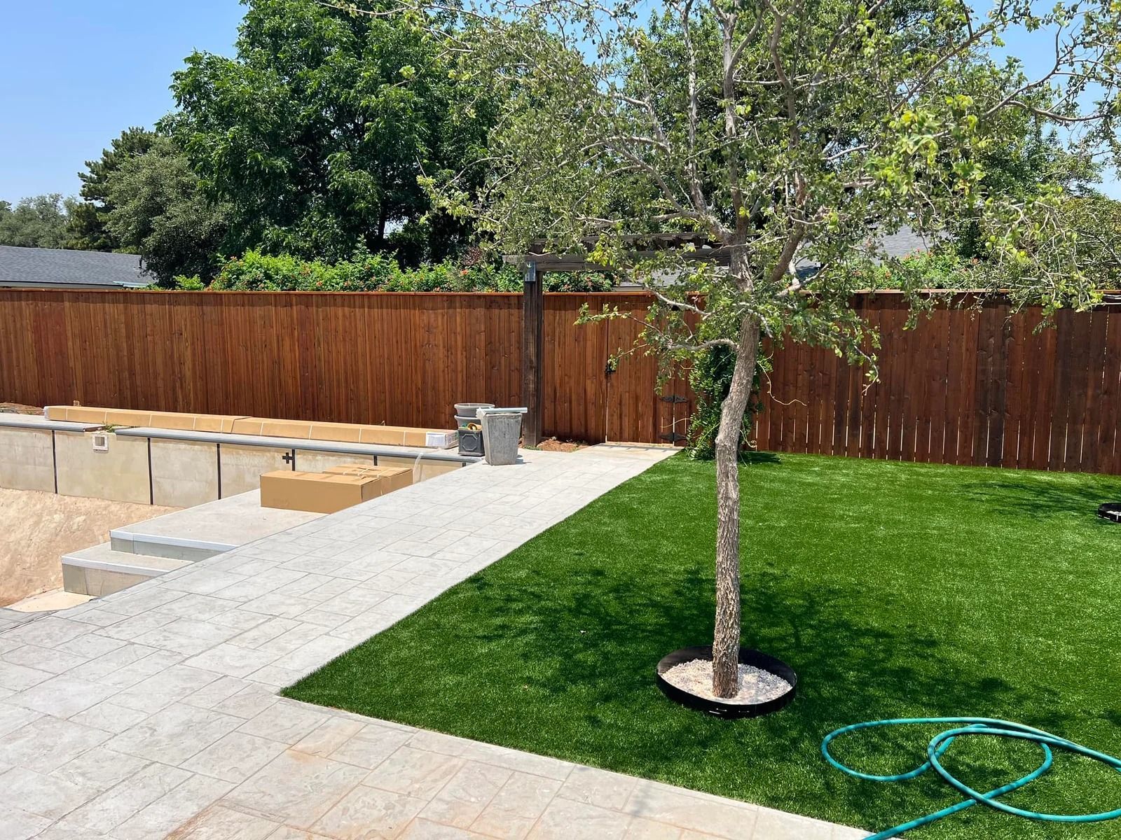 Backyard with green lawn, young tree, concrete patio, and wooden fence under a sunny sky