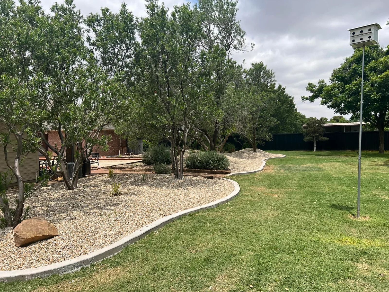 Landscaped yard with gravel beds, trees, shrubs, and a grassy lawn under a cloudy sky.