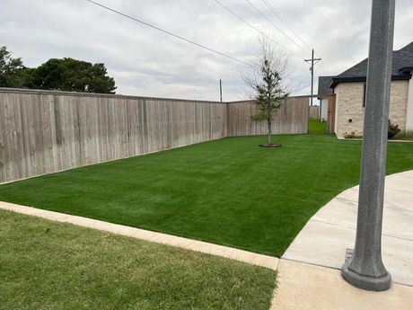 Neatly landscaped backyard with green lawn, wooden fence, young tree, and concrete path beside a house