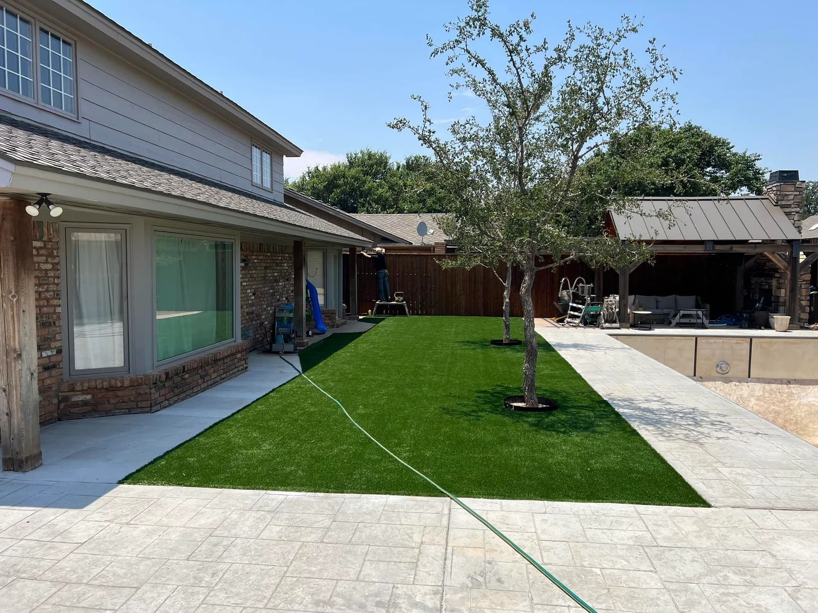 Backyard patio with a green lawn, tree, and stone paving beside a house and pool area