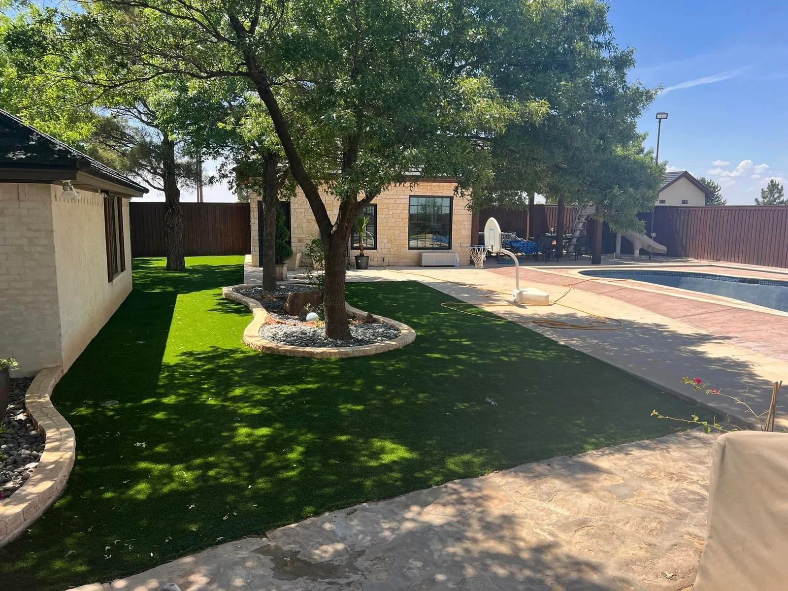 Backyard with large tree, green lawn, stone border, and a paved patio beside a house