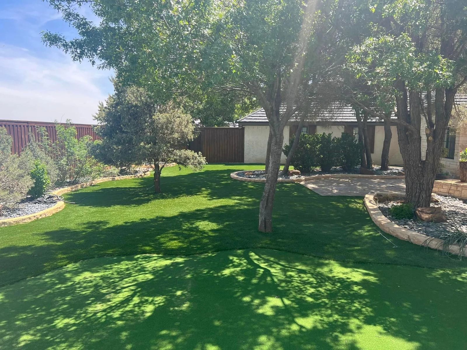 Shaded backyard with lush green lawn, trees, stone border, and a patio under a bright sky
