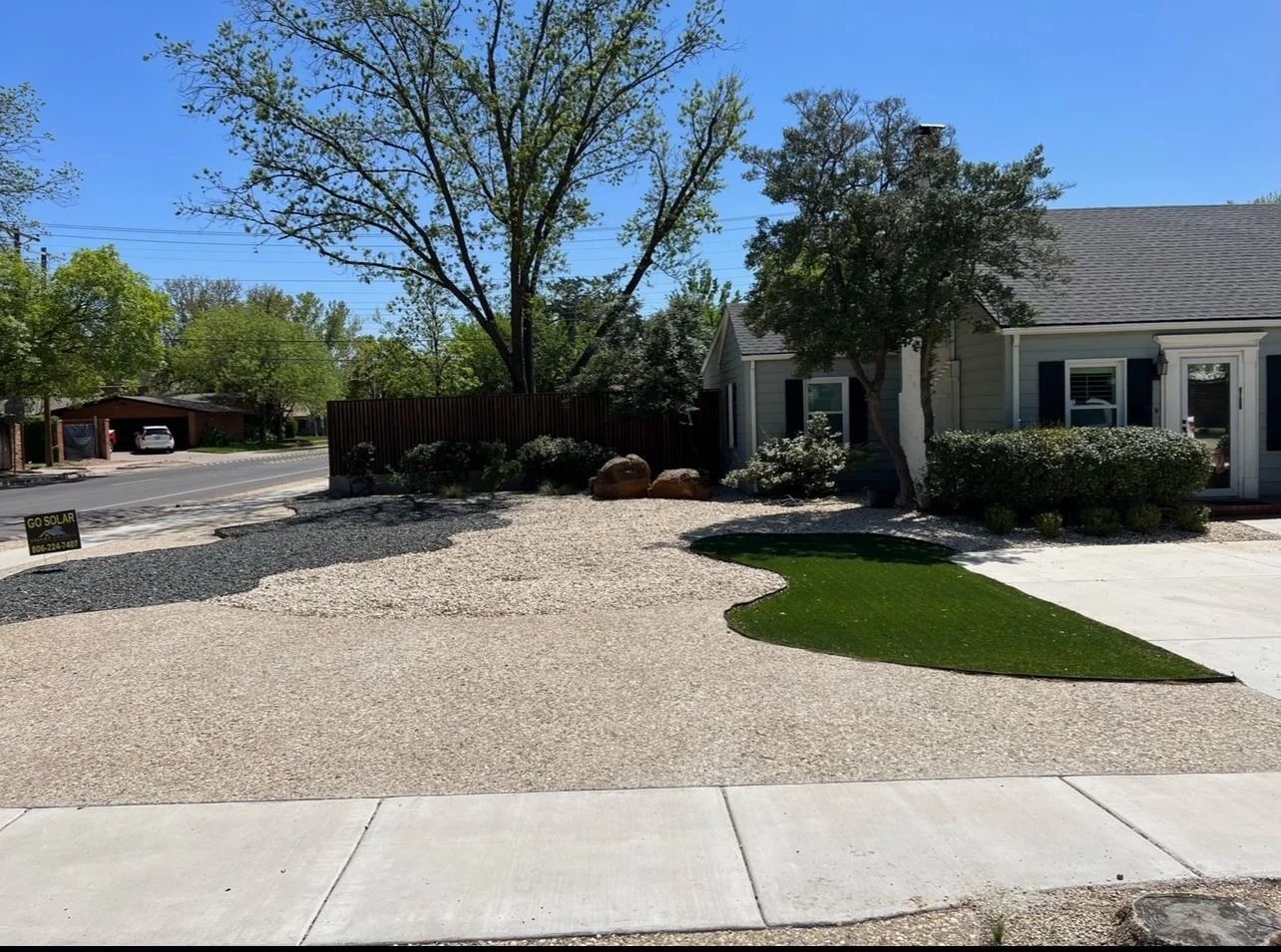 Gravel driveway and front yard beside a beige house, with trees and a small lawn under bright sunlight.