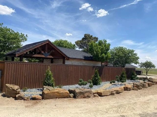 Rustic house behind a wooden fence, landscaped with rocks and shrubs under a blue sky.