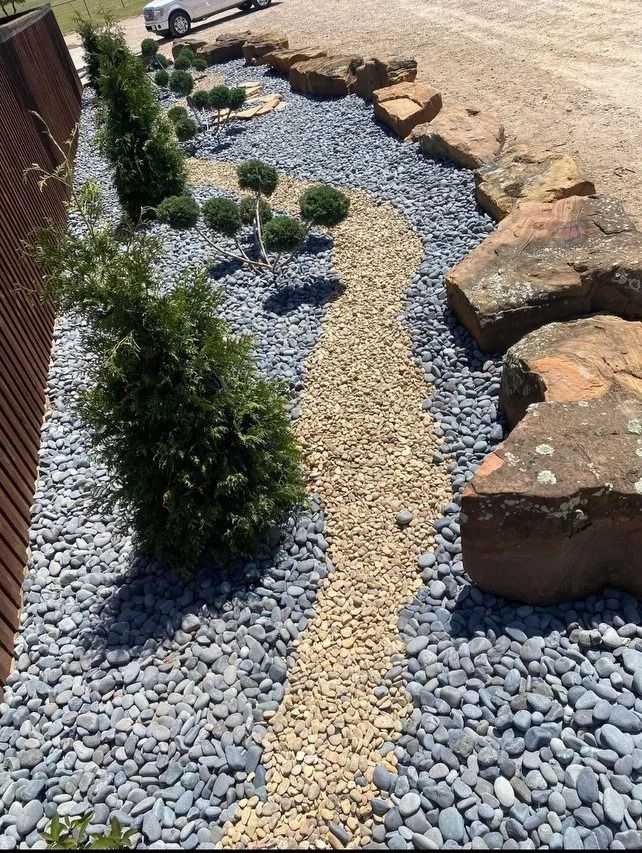 Landscaped gravel garden with shrubs, stepping stones, and large boulders beside a brick wall