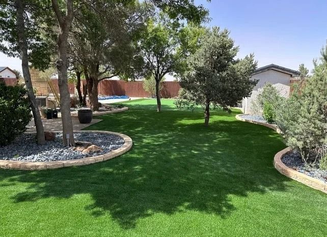 Lush green backyard with trees, a patio, and a curved stone border around the lawn