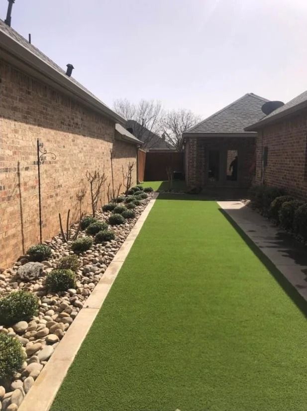 Narrow artificial turf walkway between brick walls with landscaping stones and a house in the background