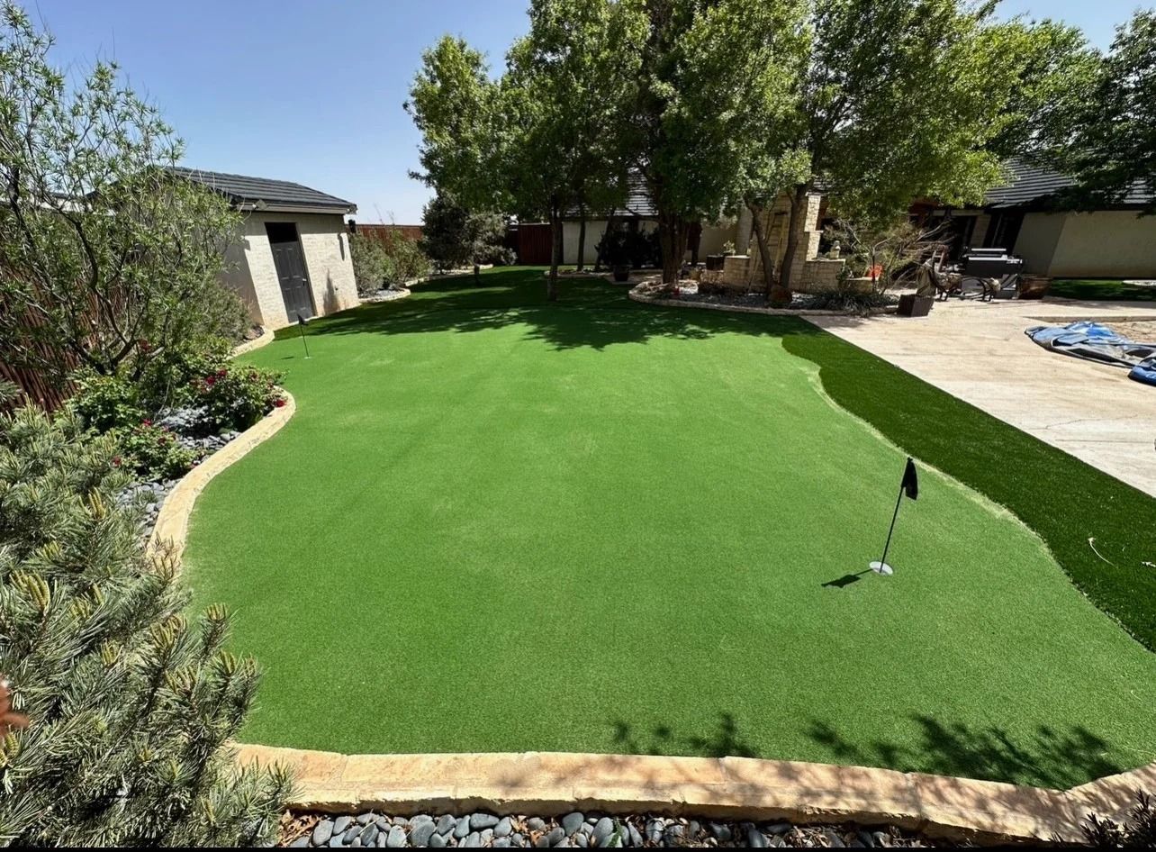 Bright green artificial turf lawn in a backyard, bordered by stone edging and trees, with a golf flag stick.