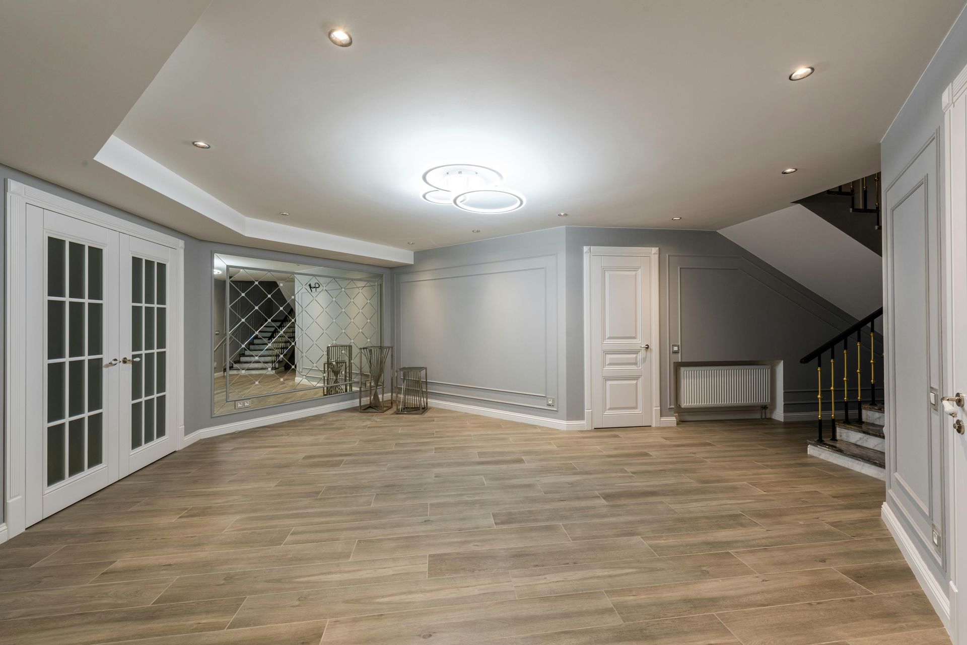 Empty modern living room with light wood floors, gray walls, French doors, and recessed ceiling lights
