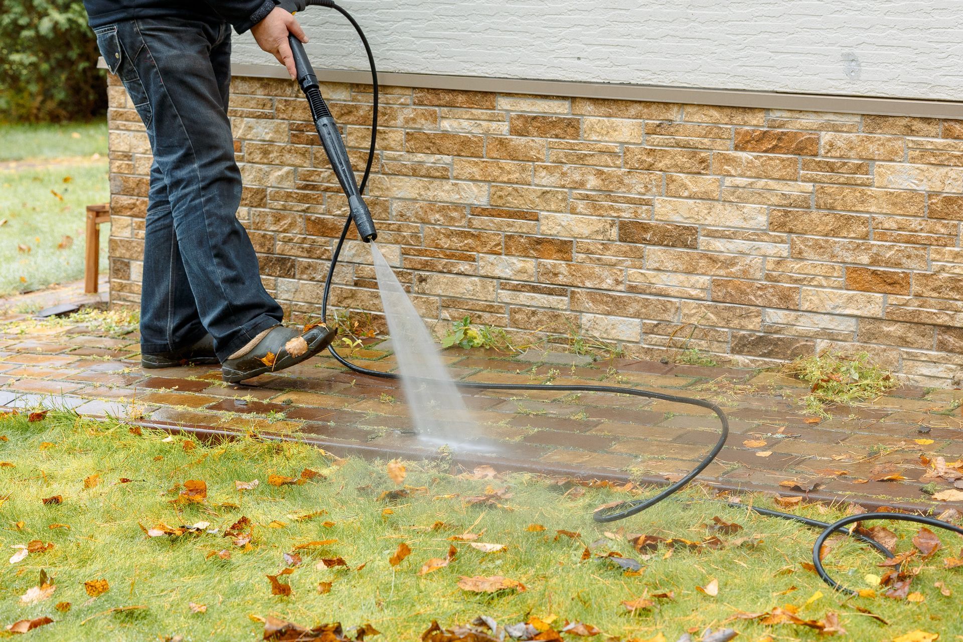 Person pressure washing a stone wall and pavement in a yard with scattered leaves
