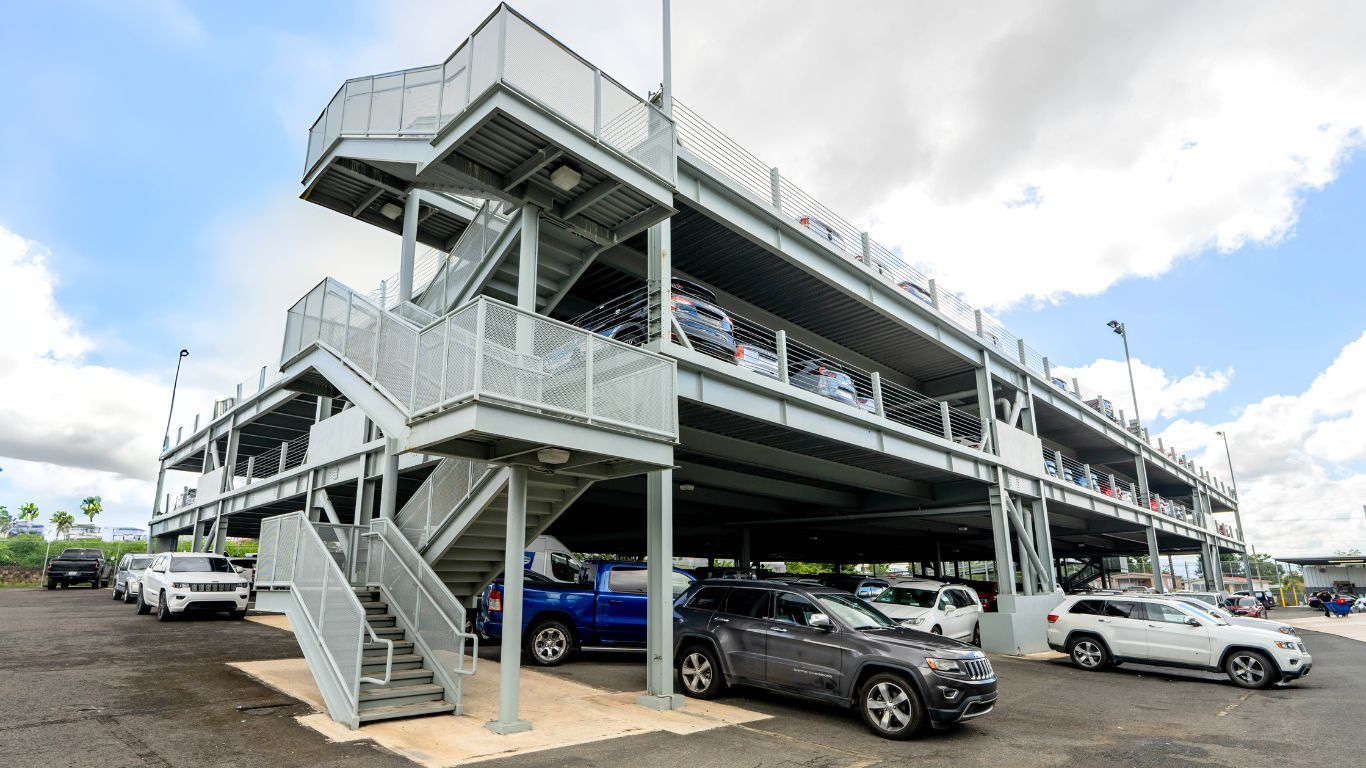 Three-story parking garage with stairwells. Cars parked on levels. Gray and white structure, blue sky.