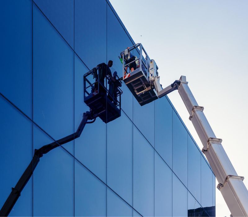 Two workers in aerial lifts cleaning or inspecting a blue glass building facade.