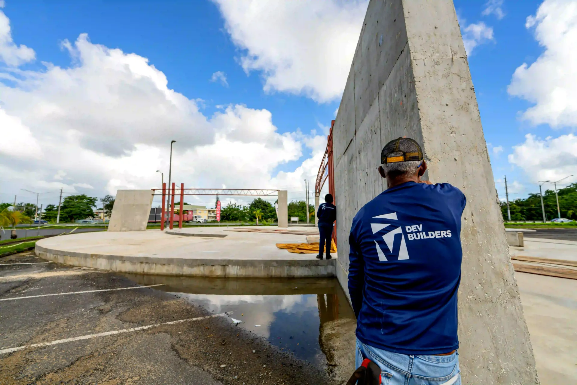 Person in blue shirt stands by a concrete wall at an outdoor construction site under a partly cloudy sky