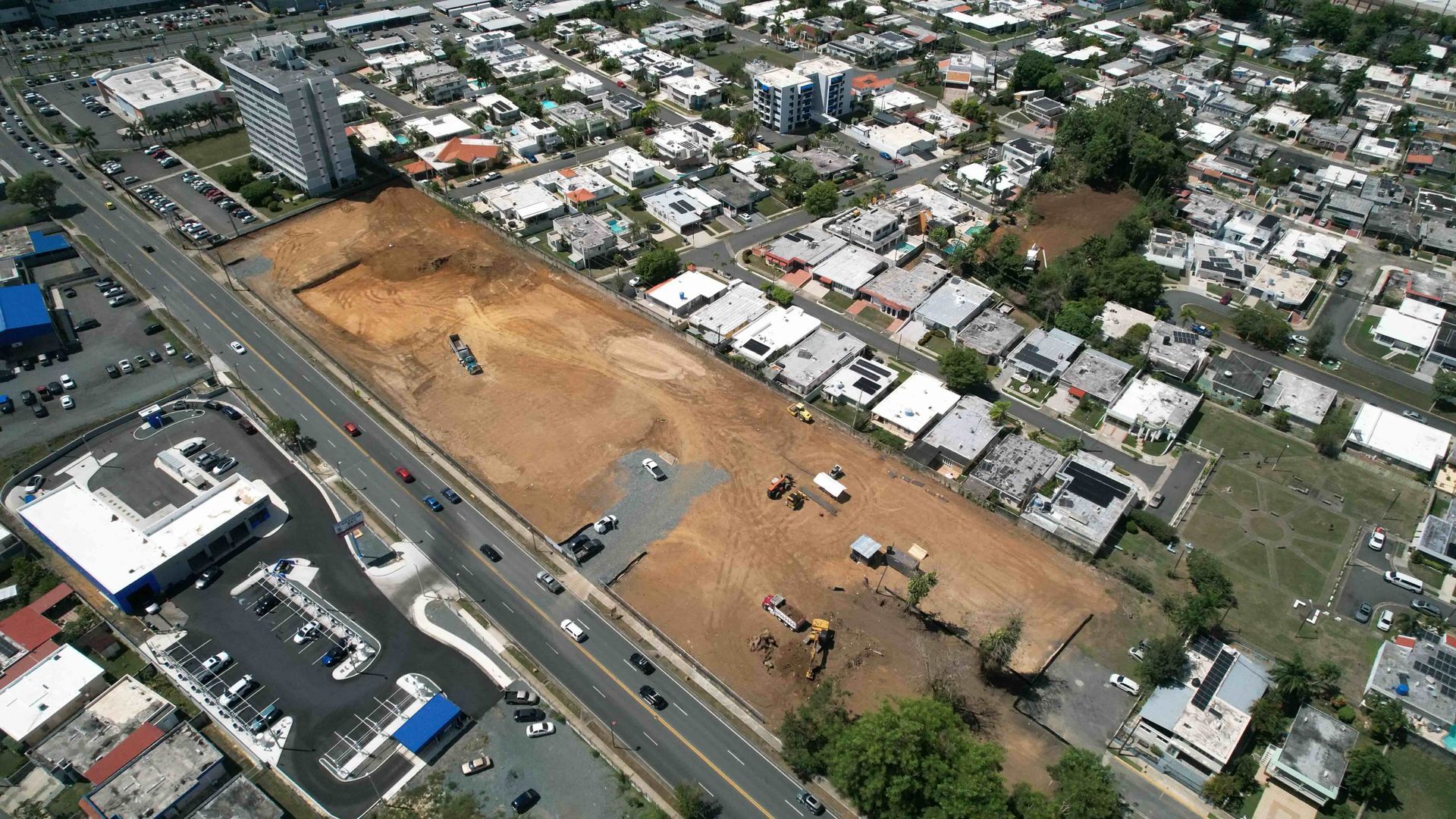 Aerial view of a large cleared dirt lot beside a road, surrounded by houses and city buildings.