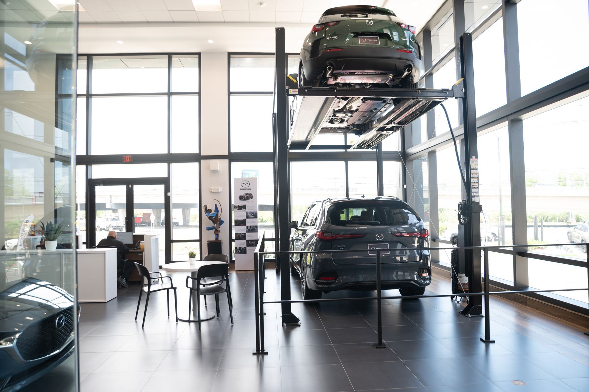 Car showroom with two SUVs, one on a car lift, one on the floor. Large windows.