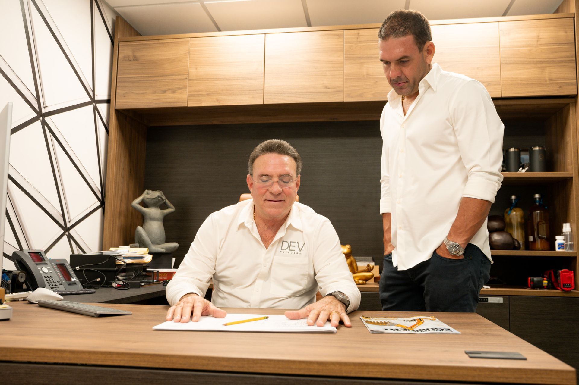 Two men in an office, one seated at a desk, the other standing, both looking at paperwork.