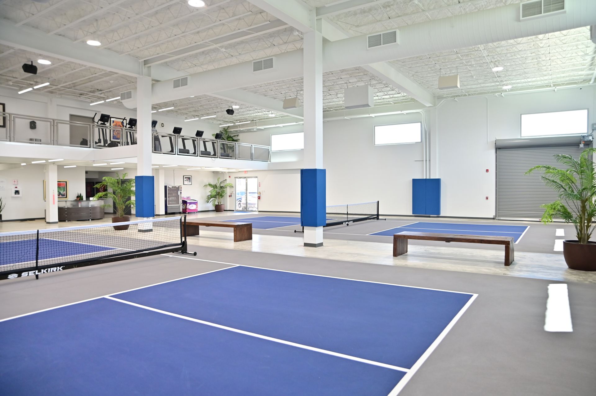 Indoor pickleball courts with blue playing surfaces, in a spacious white building with gym equipment visible.