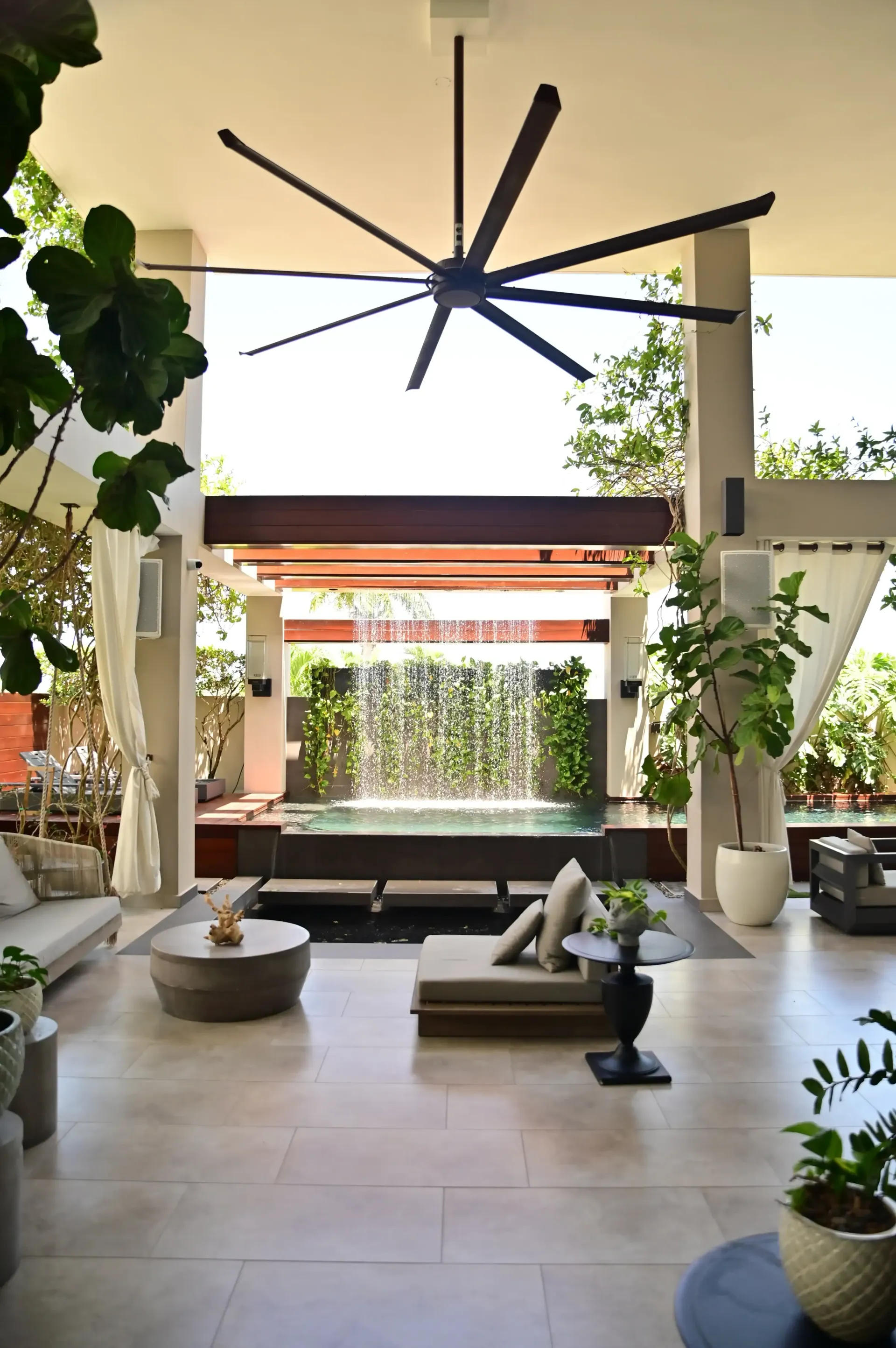 Modern hotel lobby with seating, large ceiling fan, and a water feature framed by indoor plants