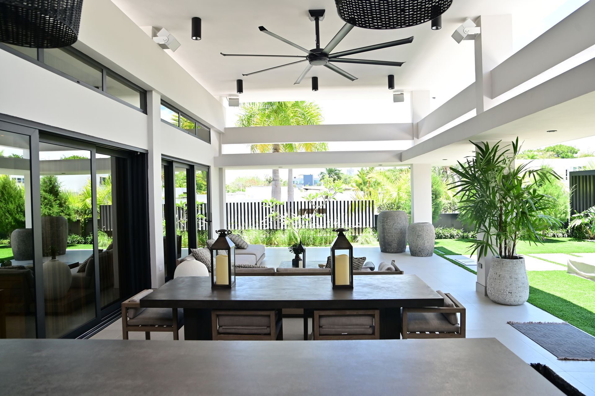 Modern patio dining area with long table, black ceiling fans, and open views to a lush garden.