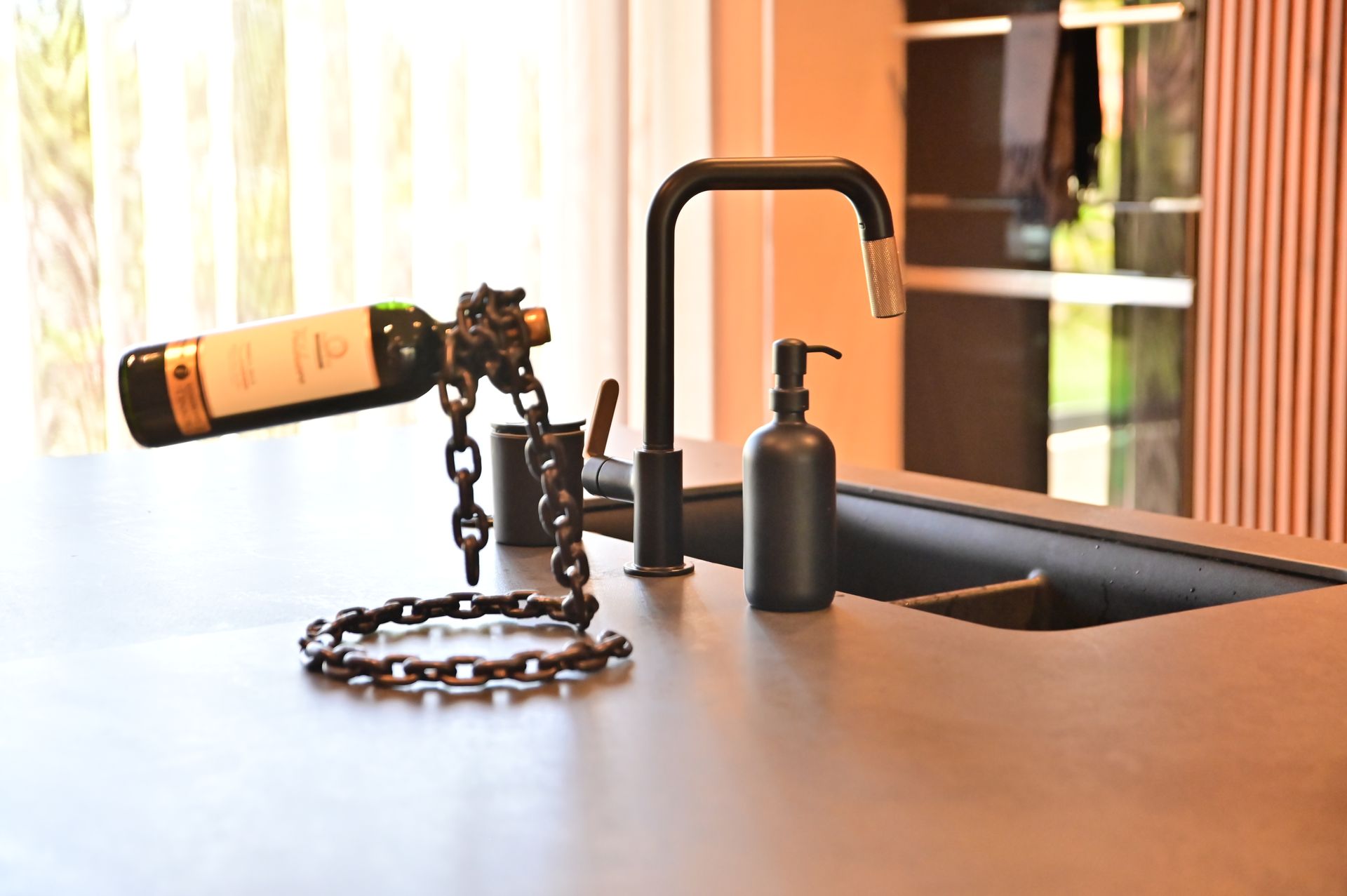 Wine bottle in a metal chain holder on a kitchen counter with a black faucet and soap dispenser.