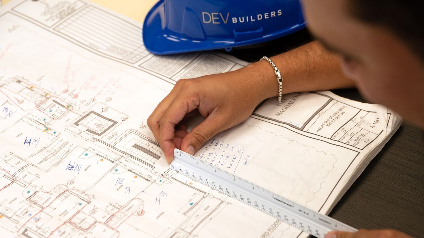 Person drafting blueprints with a ruler beside a blue hard hat on a desk