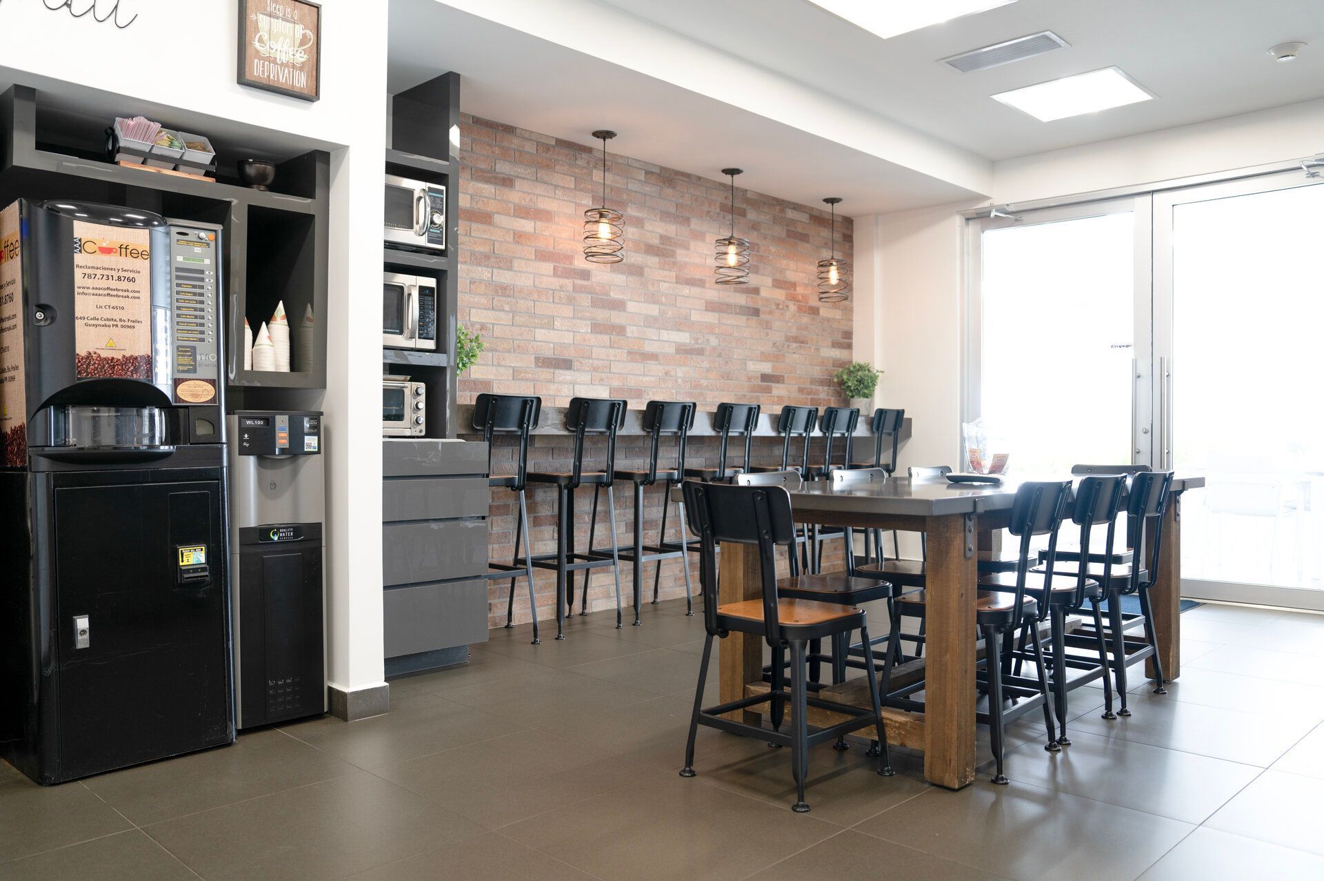 Coffee station in an office breakroom, featuring coffee machines, microwaves, high-top bar, and table with chairs.