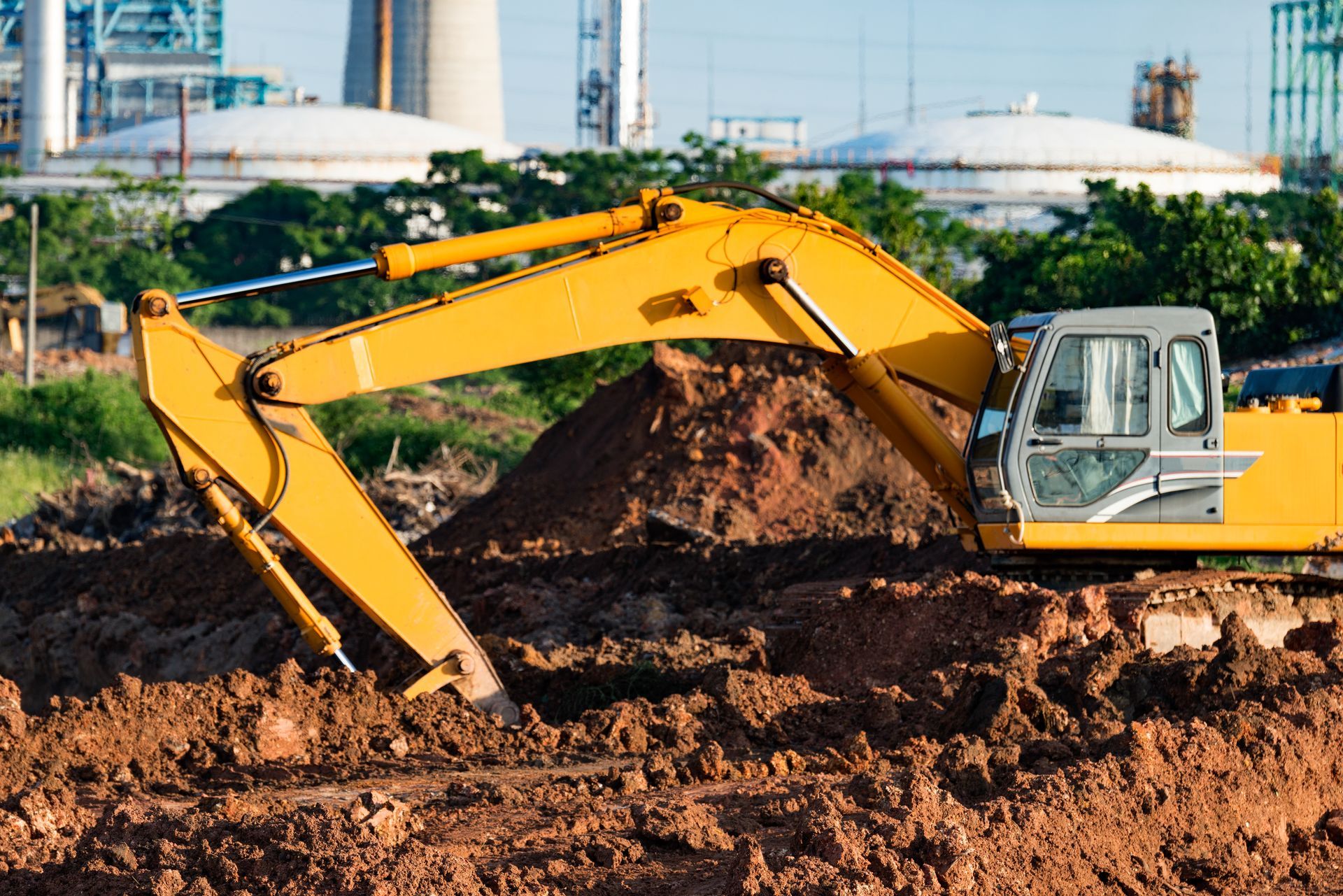 Yellow excavator digging in brown earth, with industrial buildings in the background.