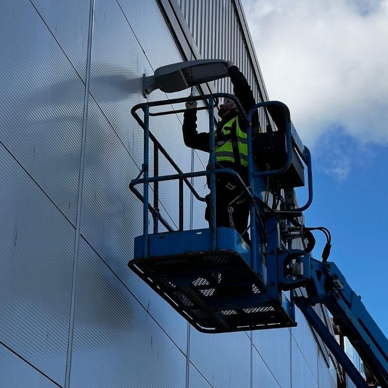 Worker on a blue lift repairs a street light on a building's metallic exterior; clear sky.