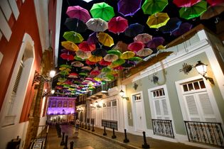 Colorful umbrellas strung overhead above a narrow, lit street at night, with white and orange buildings