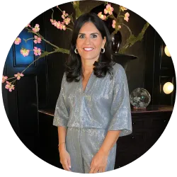 Woman in a silver dress standing indoors beside pink blossoms and a dark decorative backdrop