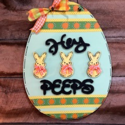 Easter egg sign with three bunny Peeps, pastel stripes, and a ribbon on a wood background