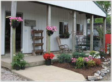 Small porch with potted flowers, a wooden chair, and a white house exterior with a garden bed