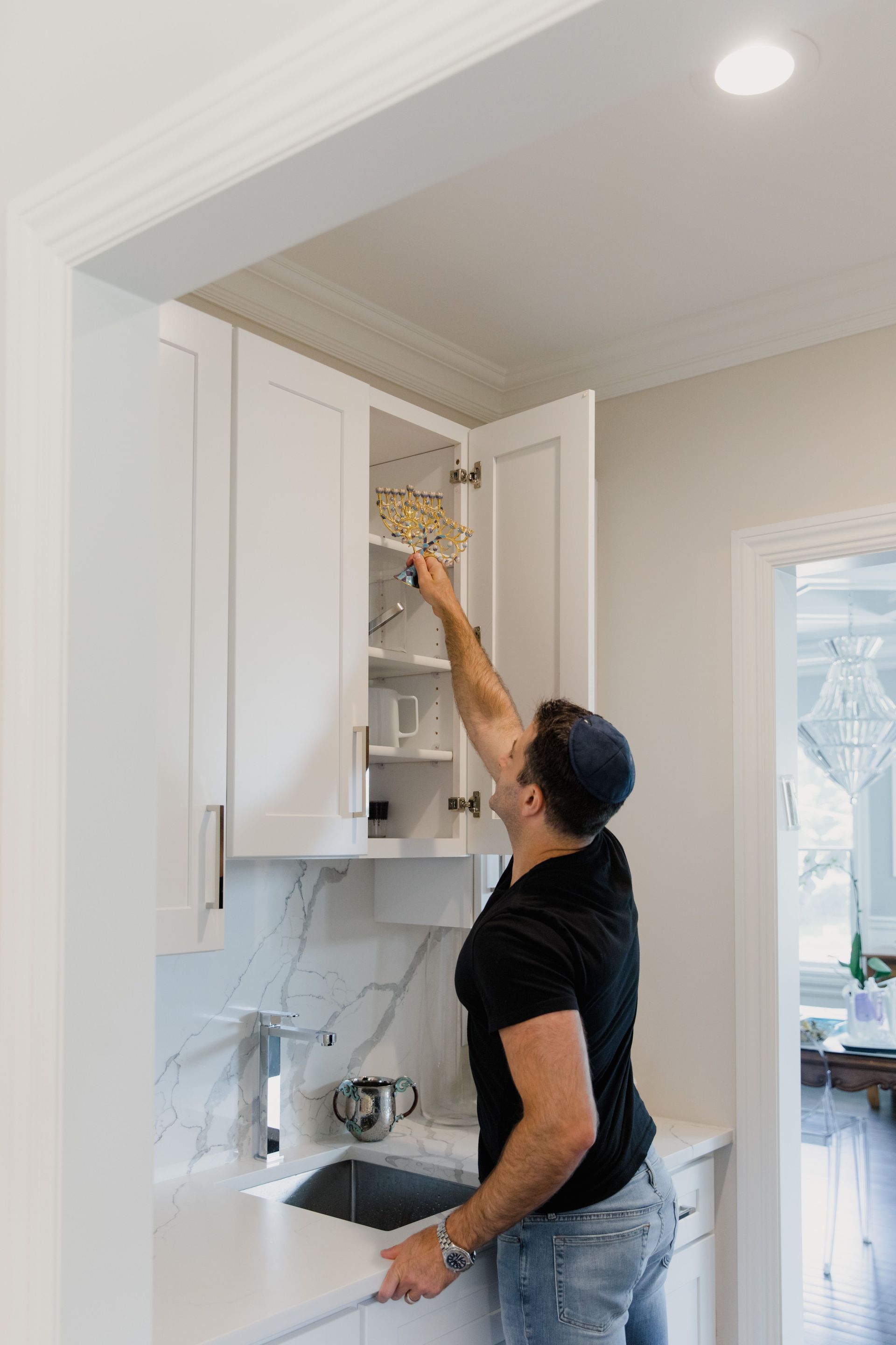 Person reaching into an open kitchen cabinet above a sink in a white kitchen