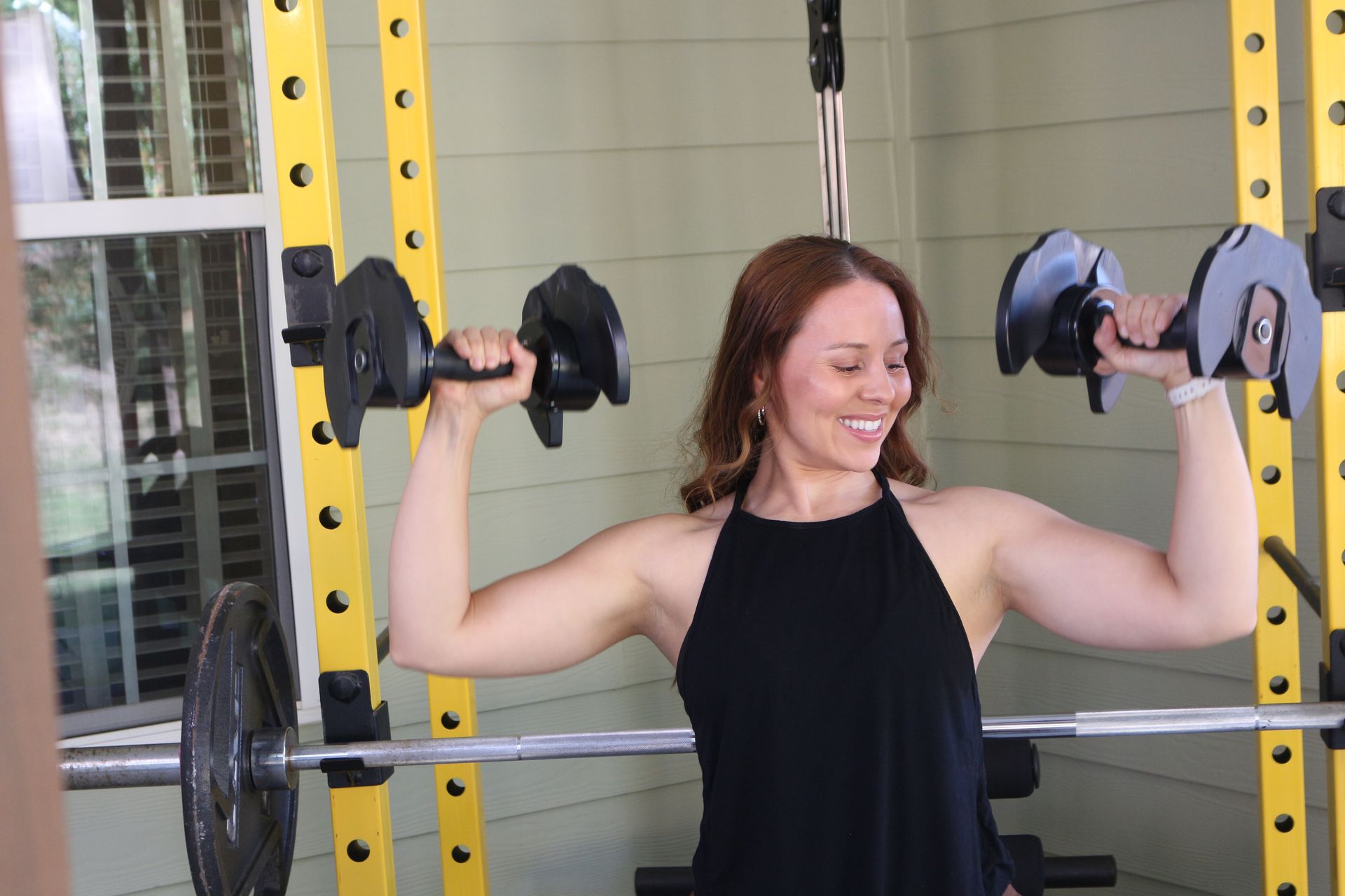Woman lifting dumbbells in home gym, smiling, with a barbell rack in background.
