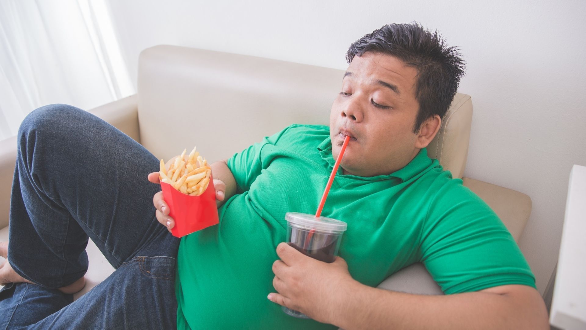 A man is laying on a couch eating french fries and drinking soda through a straw.
