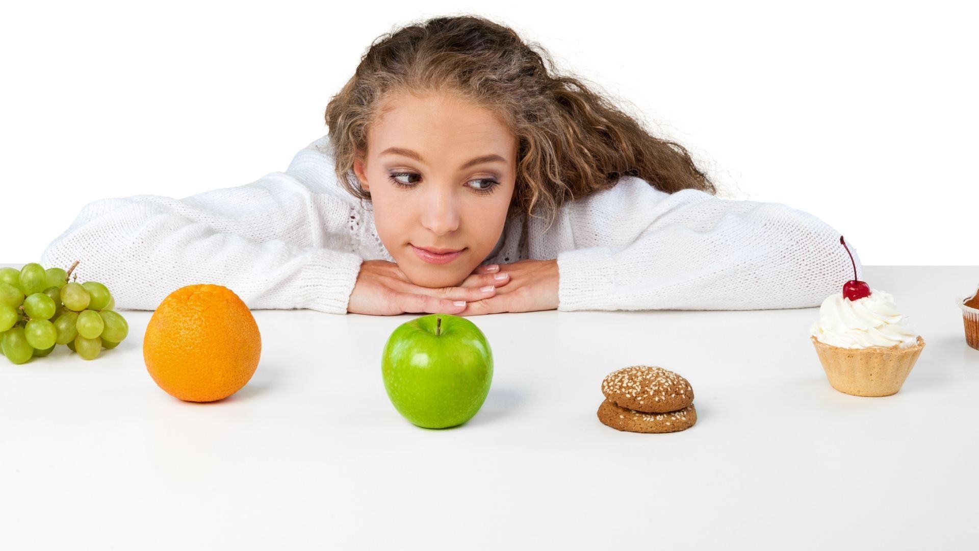 A young girl is sitting at a table with fruit , cupcakes , and a hamburger.